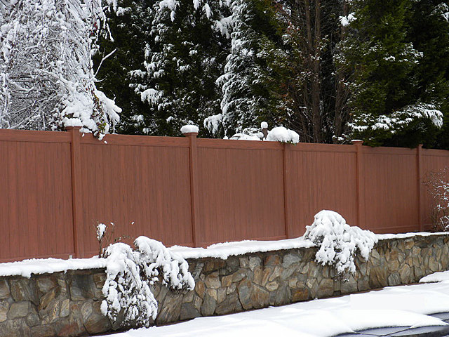 A mocha walnut colored wood grain vinyl privacy fence surrounded by snow in a yard